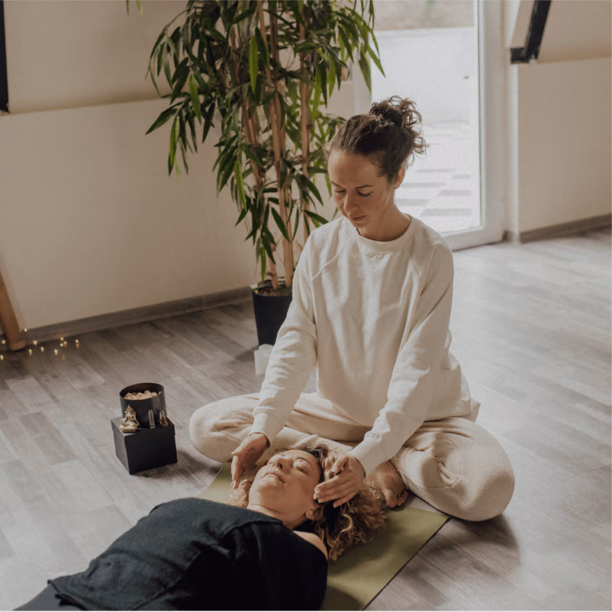 Woman in white sweater sitting cross-legged on mat giving a head massage to a woman lying down with eyes closed in a calm room with a plant and soft lighting.