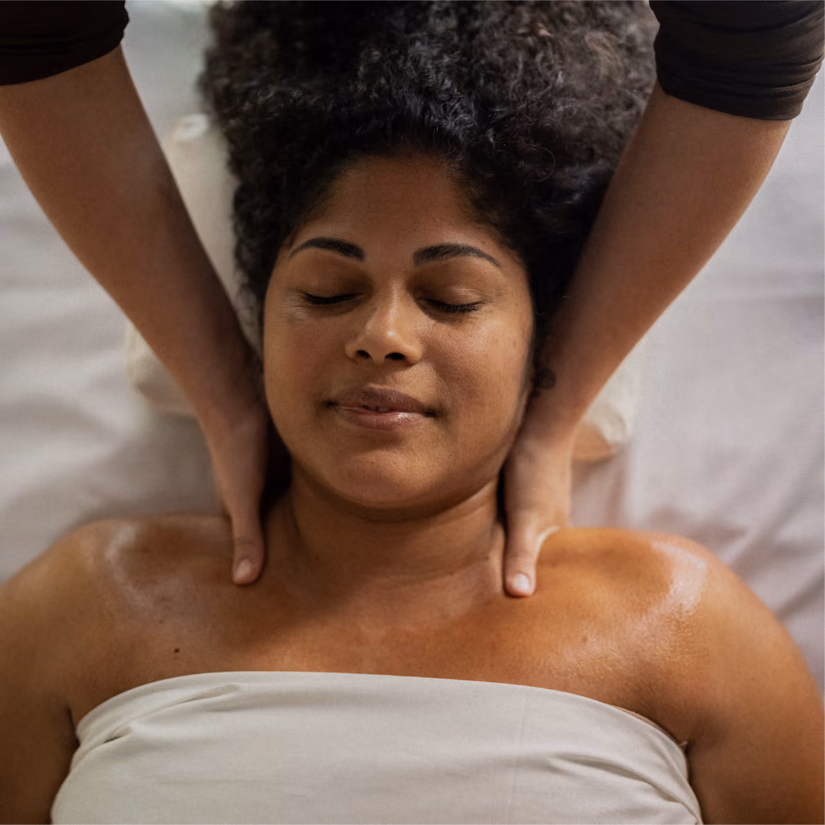 Person with eyes closed receiving a shoulder and neck massage, lying on a white sheet, appearing relaxed.