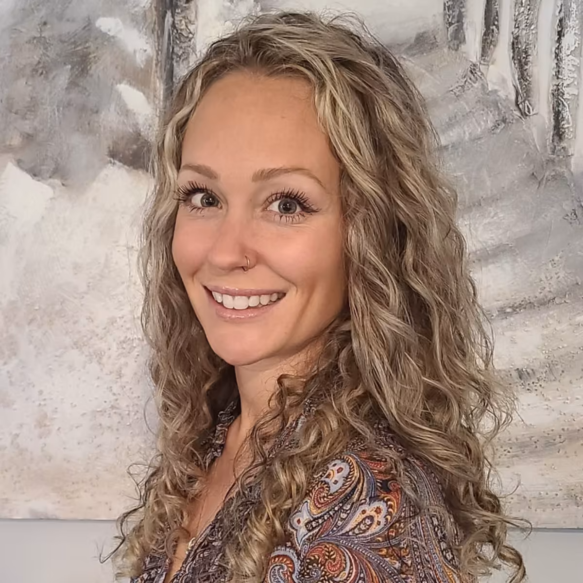 Smiling woman with long curly blonde hair, wearing a patterned top with paisley designs, posing in front of a textured gray and white wall background.