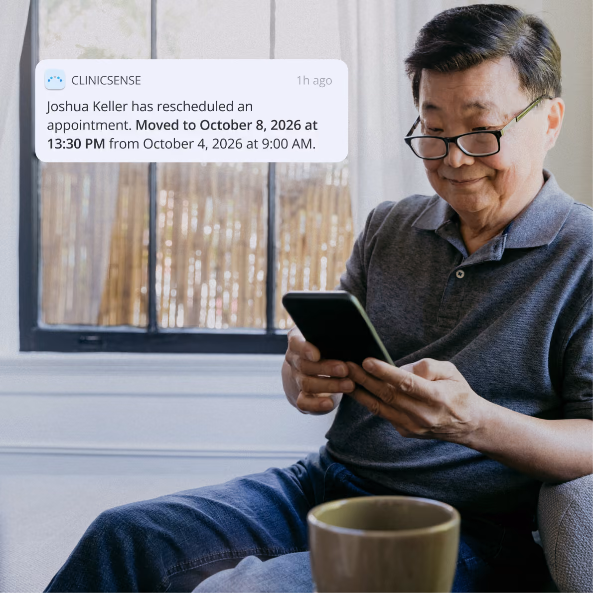 An older man with glasses, wearing a gray polo shirt and jeans, sits on a couch using a smartphone with a green cup on the table in front of him. A notification bubble appears near him, showing a message from CLINICSENSE that Joshua Keller rescheduled an appointment from October 4, 2026 at 9:00 AM to October 8, 2026 at 1:30 PM.