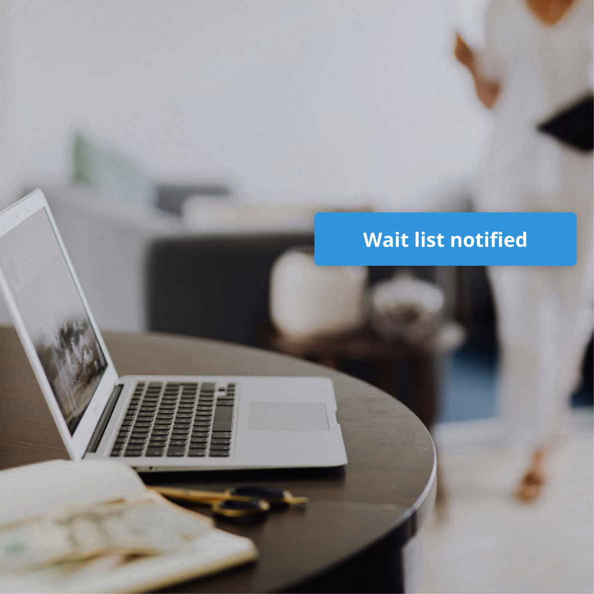 Open laptop on a round wooden table with a notebook and scissors, a blurred person holding a tablet in the background, and a blue notification bubble reading 'Appointment confirmed.'