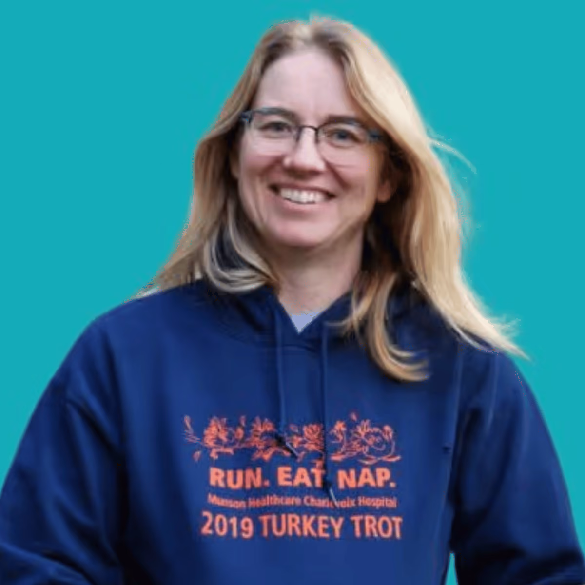 Smiling woman with blonde hair and glasses wearing a navy blue hoodie with orange text that reads 'RUN. EAT. NAP. Masson Healthcare Chase neck Hospital 2019 TURKEY TROT' on a teal background.