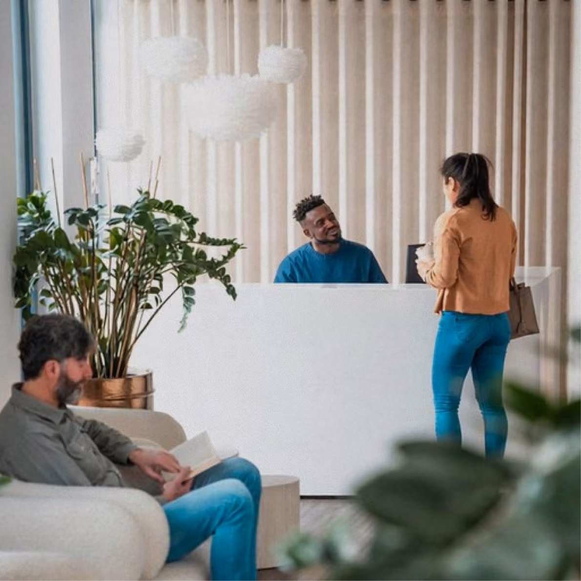 A woman stands at a white reception desk talking to a receptionist in a blue shirt, while a man with a beard sits nearby reading a book in a waiting area with plants.