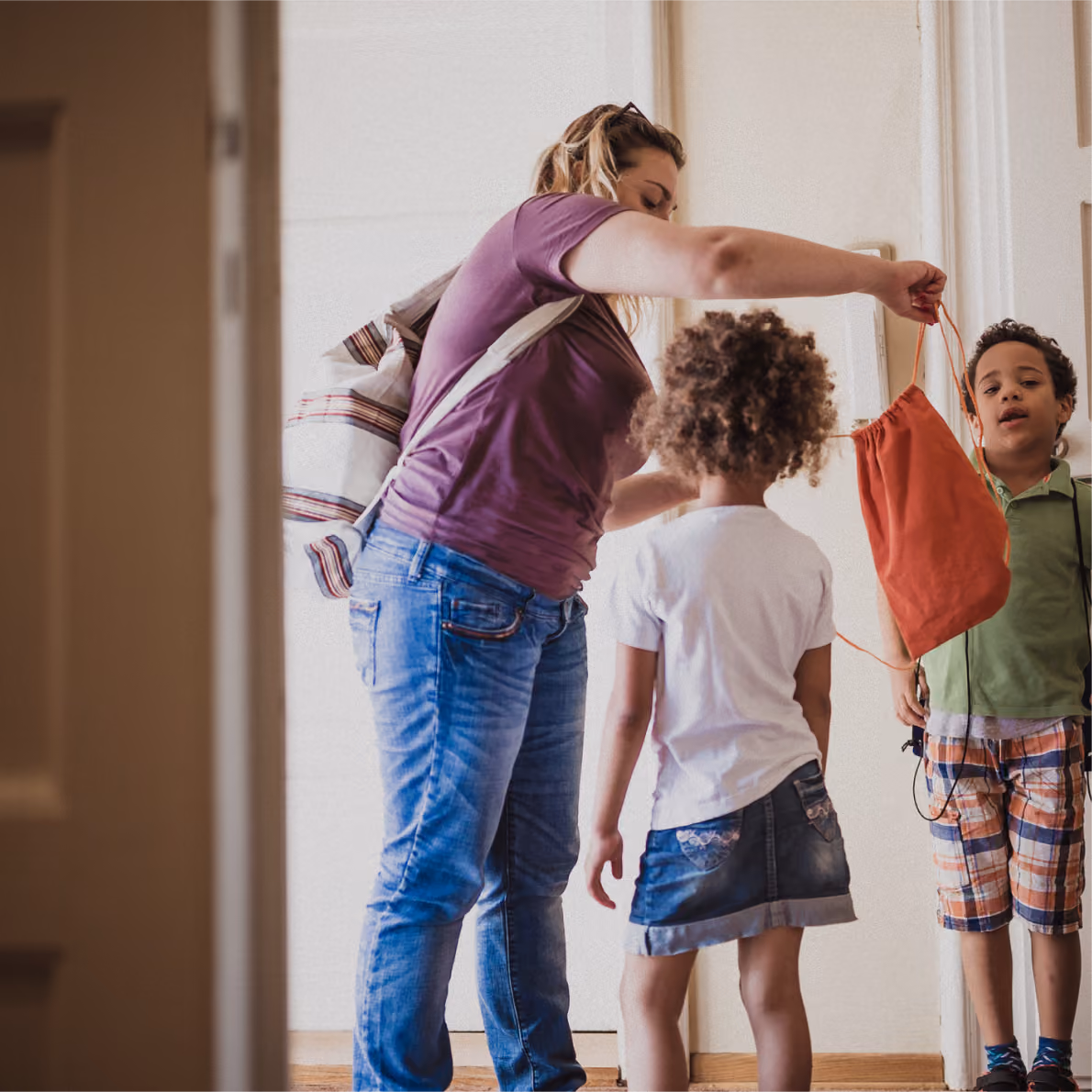 Woman holding an orange drawstring bag out to two children standing in a hallway, one child facing the woman and the other looking forward.