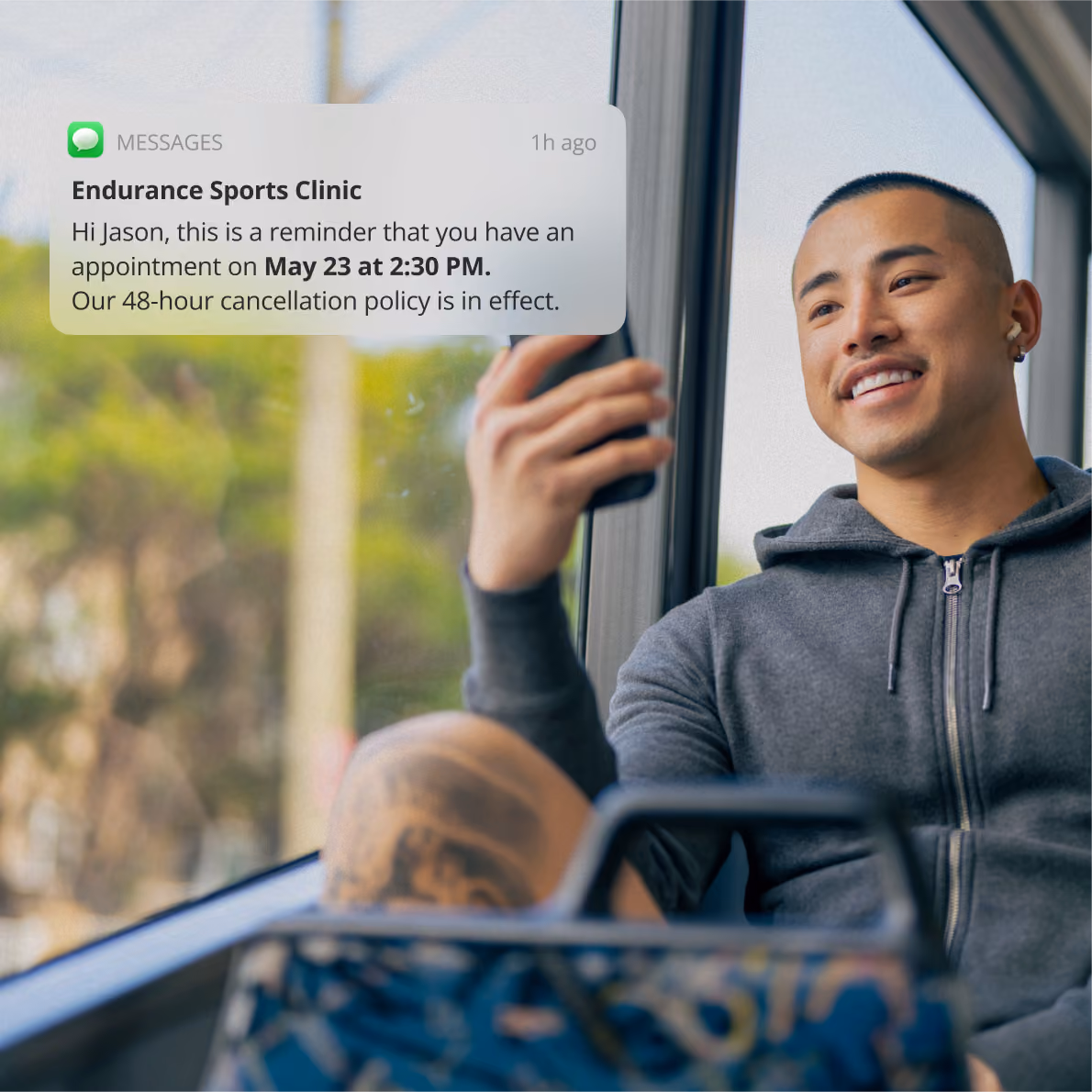Young man sitting by a window on a bus or train, smiling while looking at his smartphone, wearing a dark zip-up hoodie and earbuds, with a message bubble on screen reading 'Please complete your forms before the appointment' and a green button labeled 'Complete forms'.