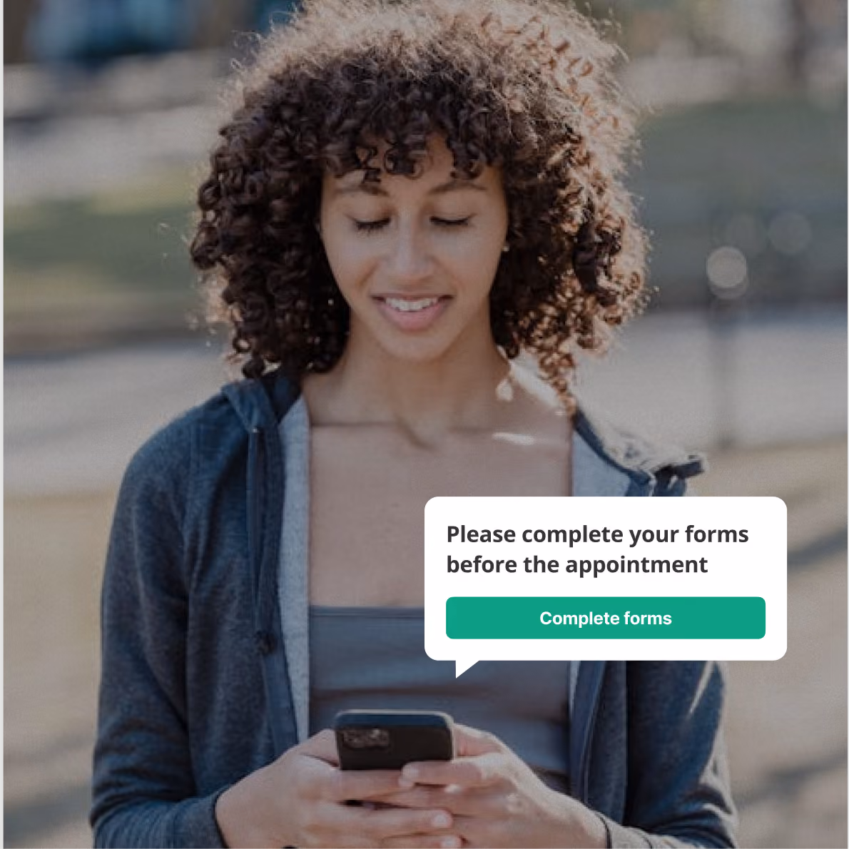 Woman with curly hair wearing a gray jacket and tank top, looking down and smiling at her smartphone, with a message bubble saying 'Please complete your forms before the appointment' and a green button labeled 'Complete forms.'