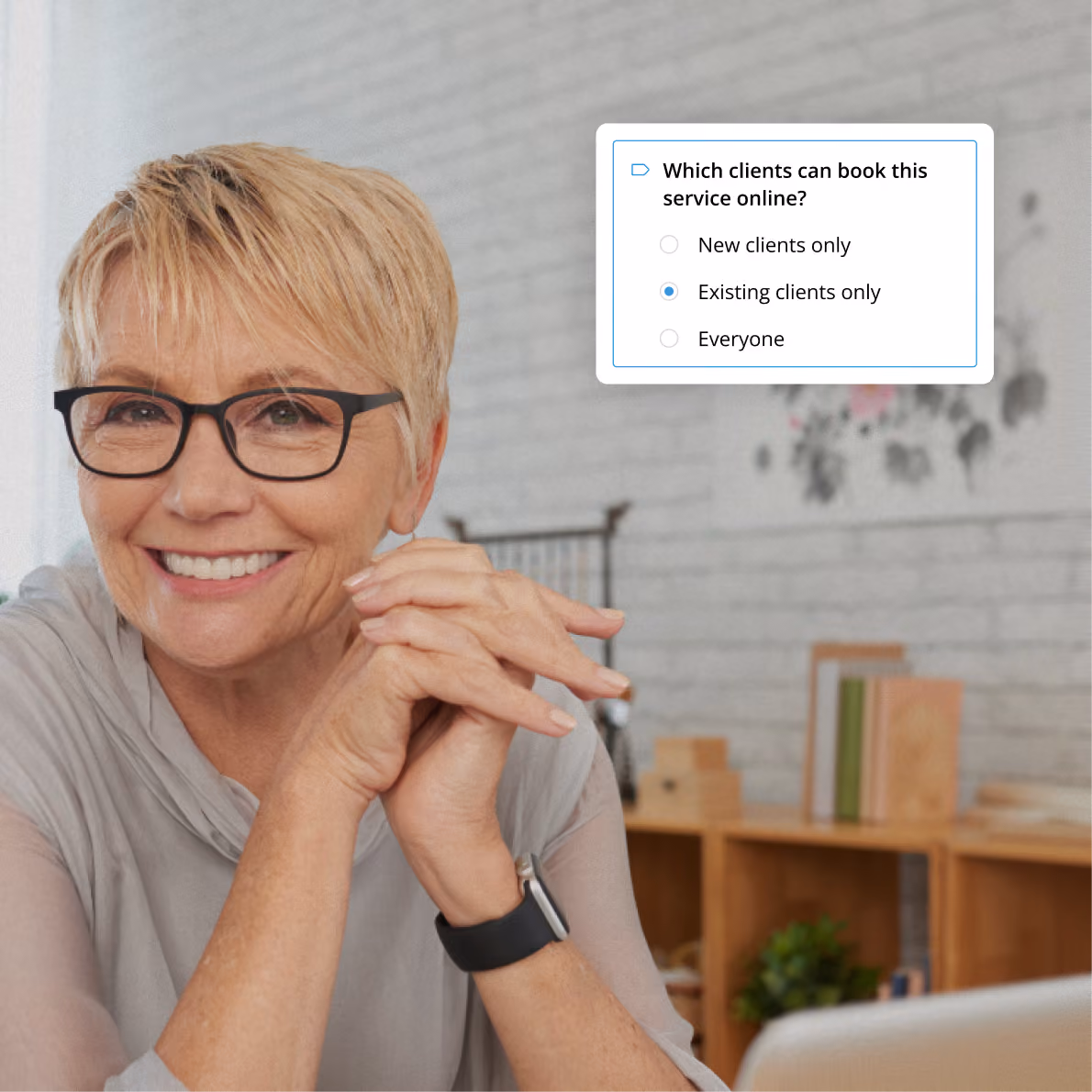 Smiling mature woman with short blonde hair and glasses seated indoors with hands clasped, next to a digital interface showing a booking setting selecting 'Existing clients only' for online service booking.