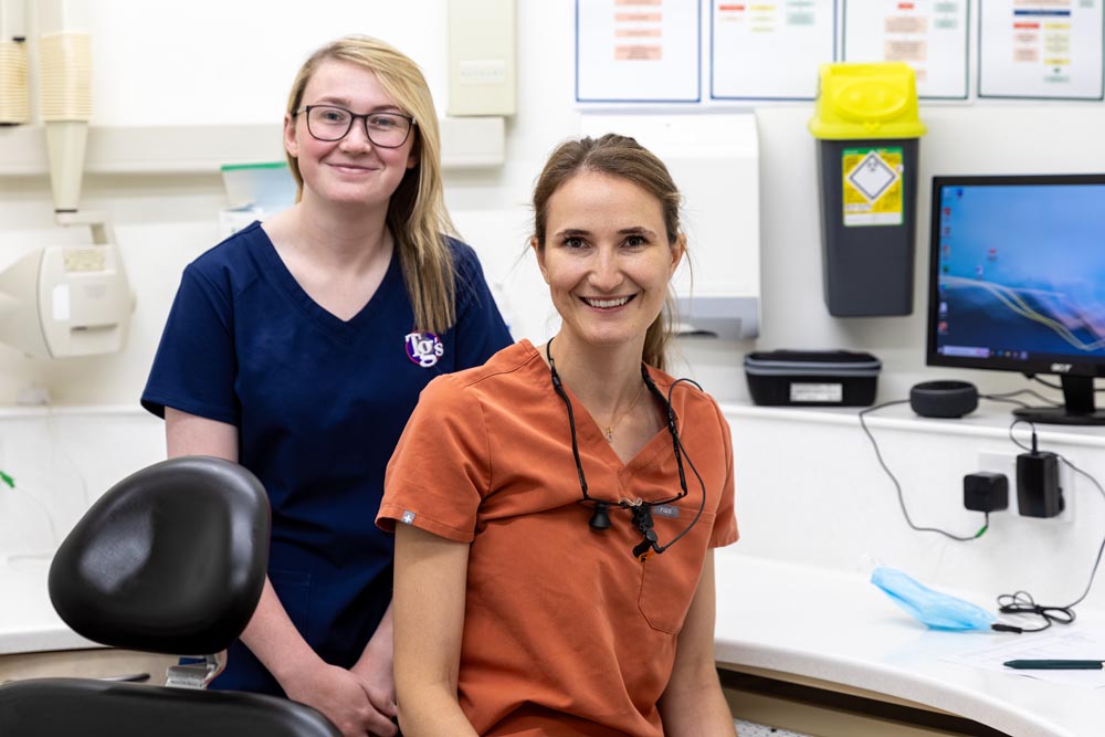 Two smiling female dental professionals in a dental office, one wearing navy scrubs and the other orange scrubs with dental loupes around her neck.