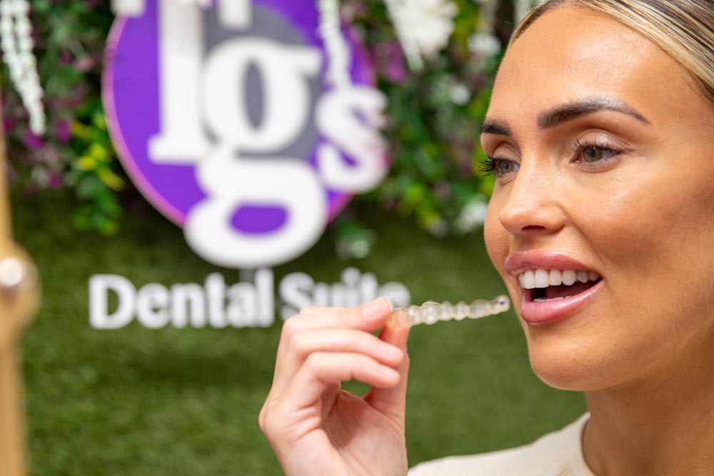 Woman smiling while holding a clear dental aligner near her mouth in front of a green wall with a blurred 'TGS Dental Suite' sign.