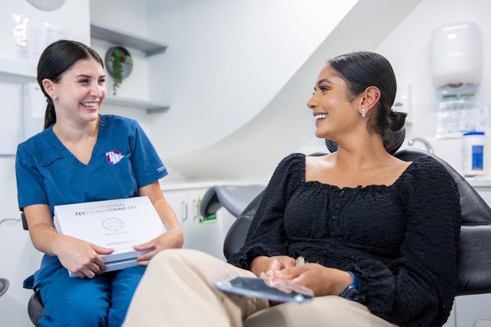 Dental professional in blue scrubs smiling and holding a teeth whitening gel box while talking to a seated patient in a dental office.