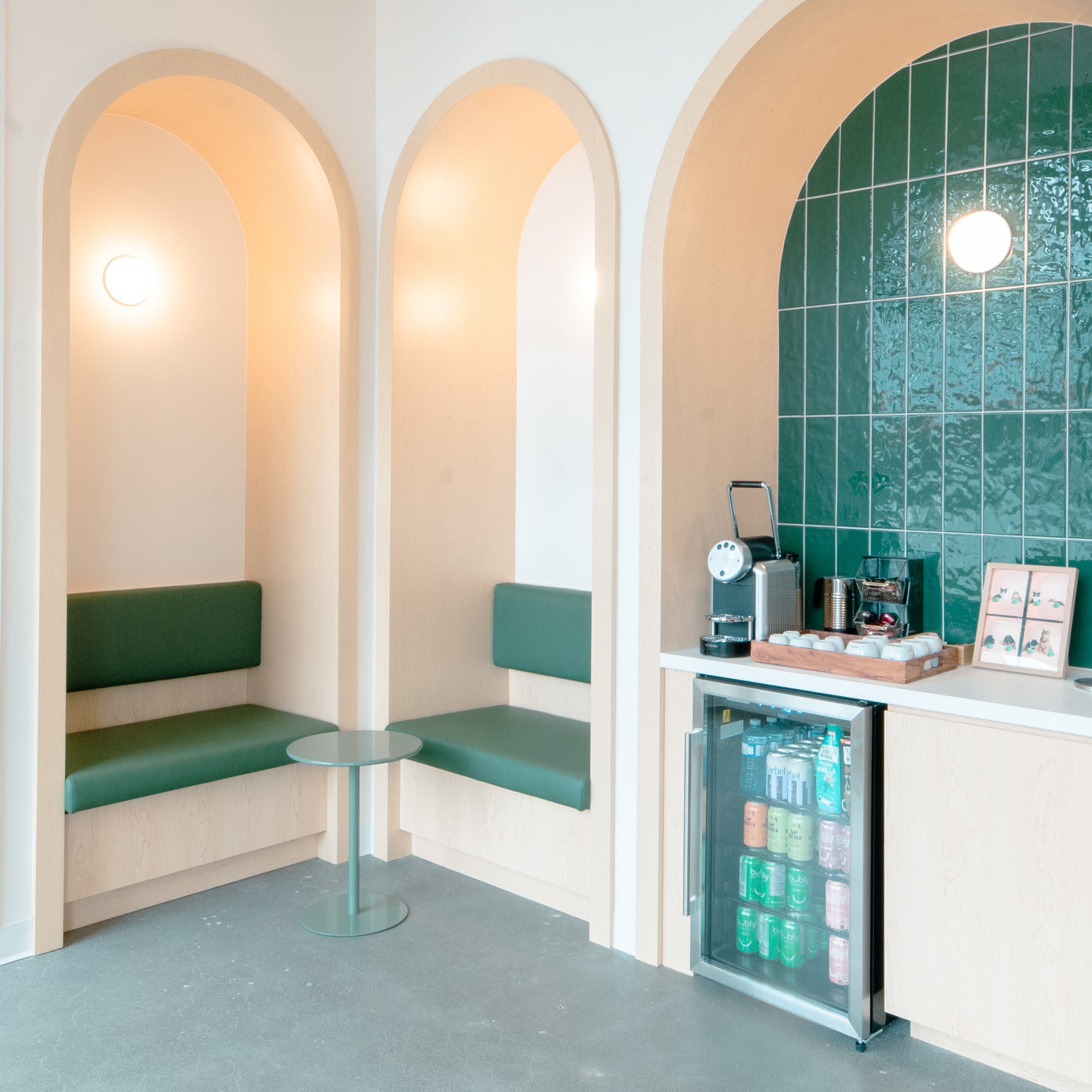 Modern veterinary clinic waiting area with three arched alcoves featuring green seating, a small table, and a refreshment station with coffee machine, refrigerator, and green tiled wall.
