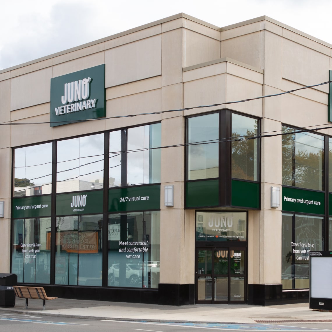 Exterior of Juno Veterinary’s Eglinton location, featuring a modern beige and glass corner building with green signage displaying “Juno Veterinary,” “Primary and urgent care,” and “24/7 virtual care,” situated along a city street with benches and power lines visible.