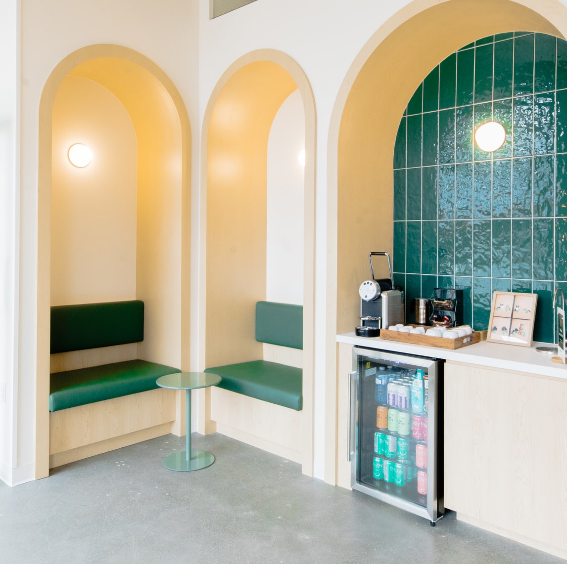 A modern lobby featuring arched wooden seating nooks with green cushions and a refreshment station with a green tiled backsplash.