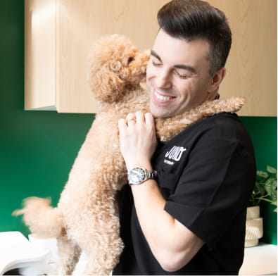 Dr. Adam, from our Main Street clinic, in black scrubs smiling as a small curly poodle licks his cheek in a clinic.