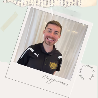 Man smiling wearing a black Puma sports shirt with a gold logo patch, standing in front of light-colored curtains. The photo is styled like a Polaroid with the word 'Happiness' handwritten below the image and a circular text 'The Whitening Clinic' partially visible.