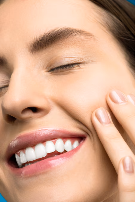 Close-up of a smiling woman with eyes closed touching her cheek, showing white teeth and natural makeup.