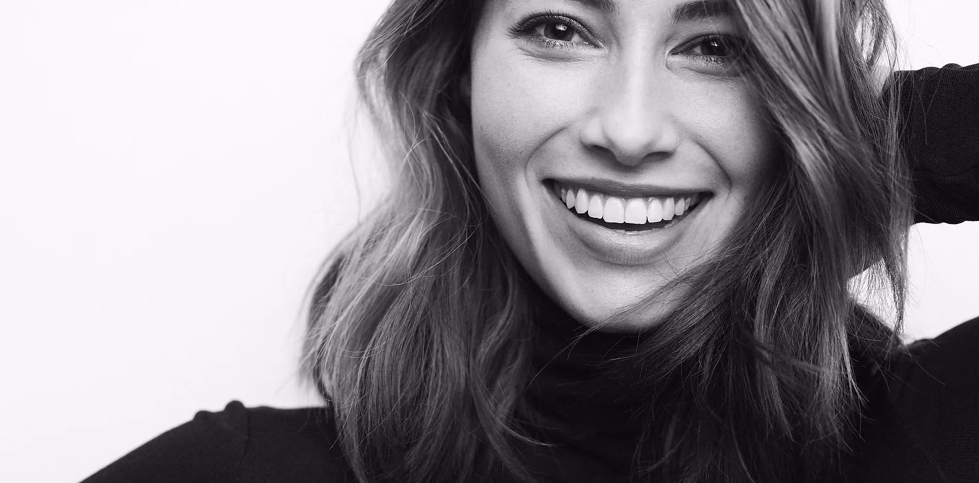 Black-and-white close-up of a smiling woman with shoulder-length hair wearing a black turtleneck, looking at the camera with one hand behind her head.