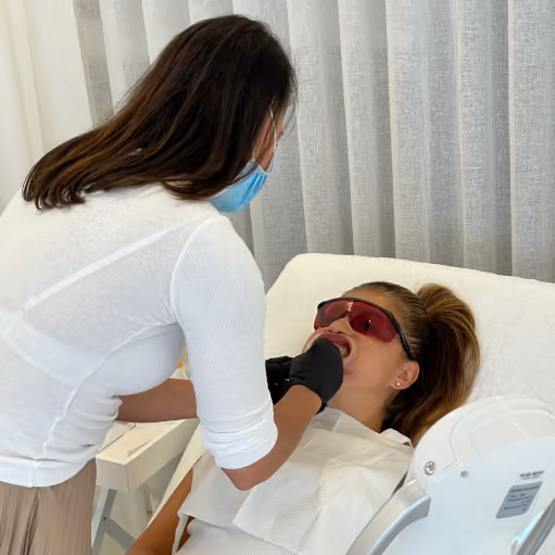 A dental professional wearing a blue face mask and white top is examining a patient's mouth; the patient reclines on a dental chair wearing protective red glasses and a dental bib.