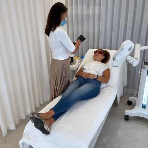 Woman in sunglasses lying on a white treatment bed covered with a towel in a medical or cosmetic clinic, while another woman wearing gloves and a face mask stands next to her preparing for a procedure with medical equipment nearby.