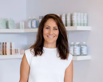 Smiling woman with long brown hair wearing a sleeveless white top standing in front of shelves with skincare products in a bright room.