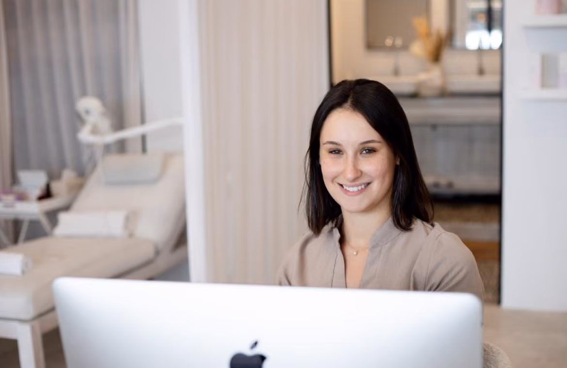 Smiling woman with dark hair sitting and looking at an Apple computer screen in a bright, modern room with a lounge chair and sink in the background.