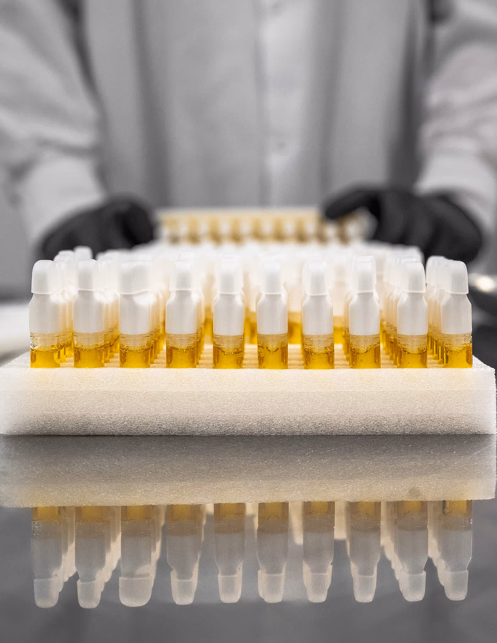 Rows of vape cartridges filled with golden liquid on a white foam tray with a person wearing a white coat and black gloves in the background.