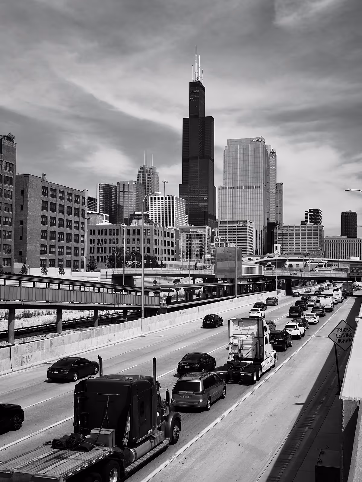 Highway traffic with trucks and cars heading towards a city skyline featuring the Willis Tower.