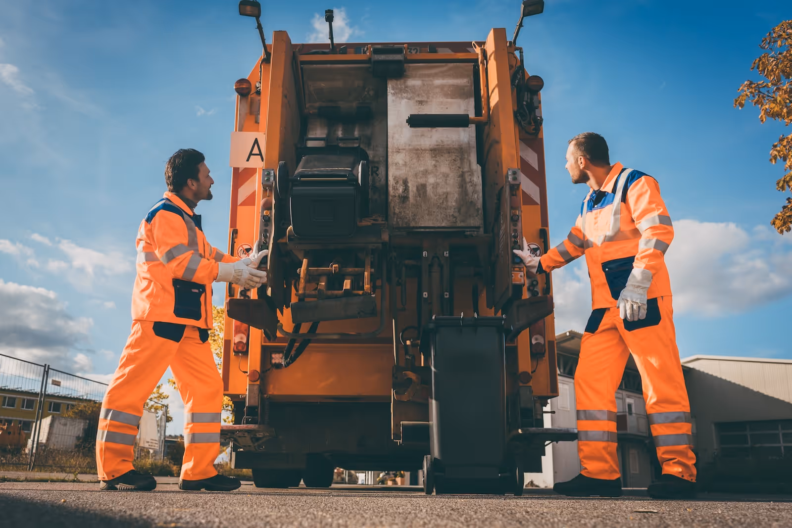 Zwei Müllwerker in orangefarbenen, reflektierenden Schutzuniformen bedienen ein Müllfahrzeug, um eine schwarze Mülltonne zu entleeren.