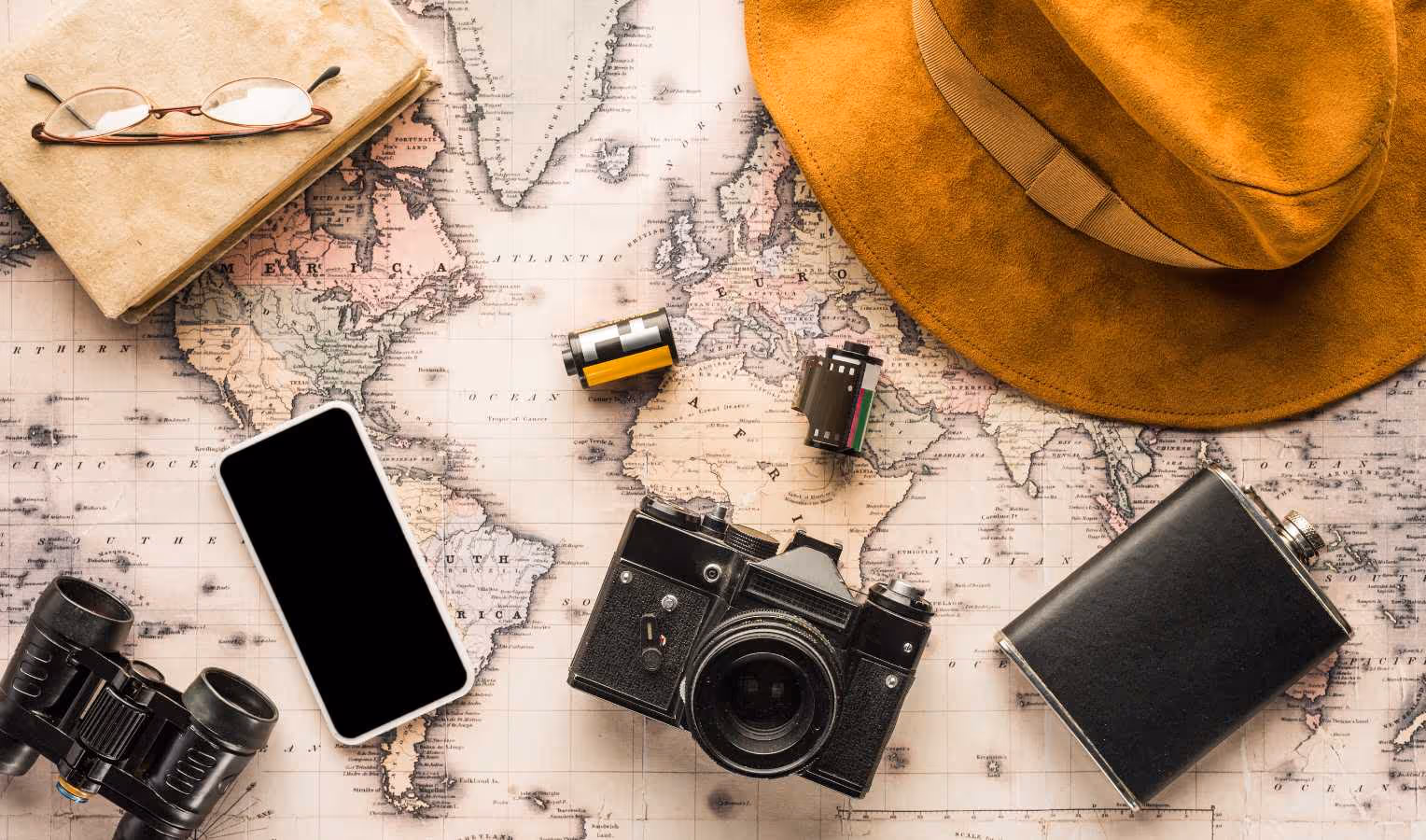 Flatlay: Reisestillleben auf alter Weltkarte mit braunem Hut, analoger Kamera, zwei Filmrollen, Smartphone, Feldflasche, Fernglas sowie Notizbuch mit Brille.
