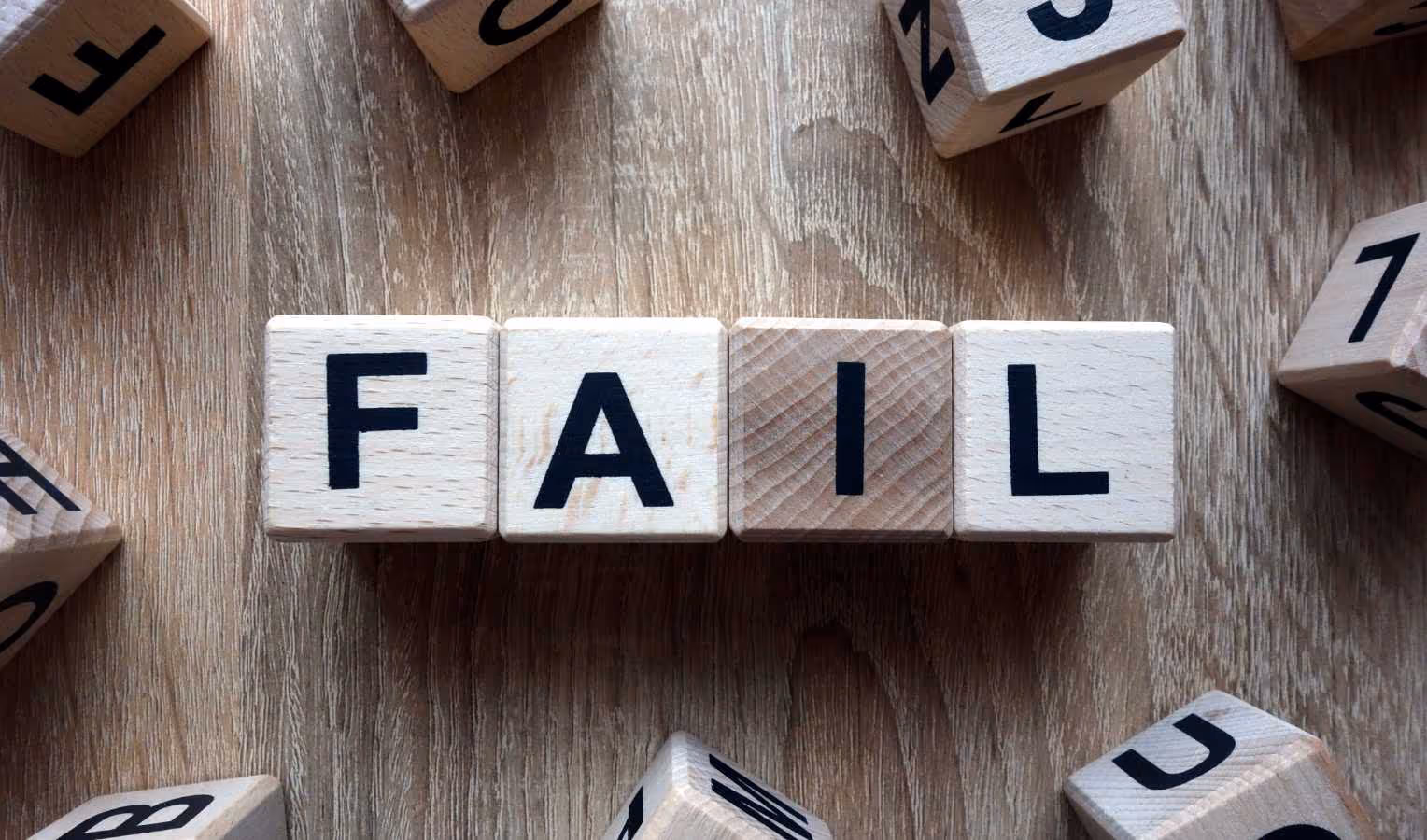 Nahaufnahme von Holzbuchstaben-Würfeln, die auf einem Holztisch das Wort „FAIL“ bilden; weitere verstreute Würfel liegen rundherum.