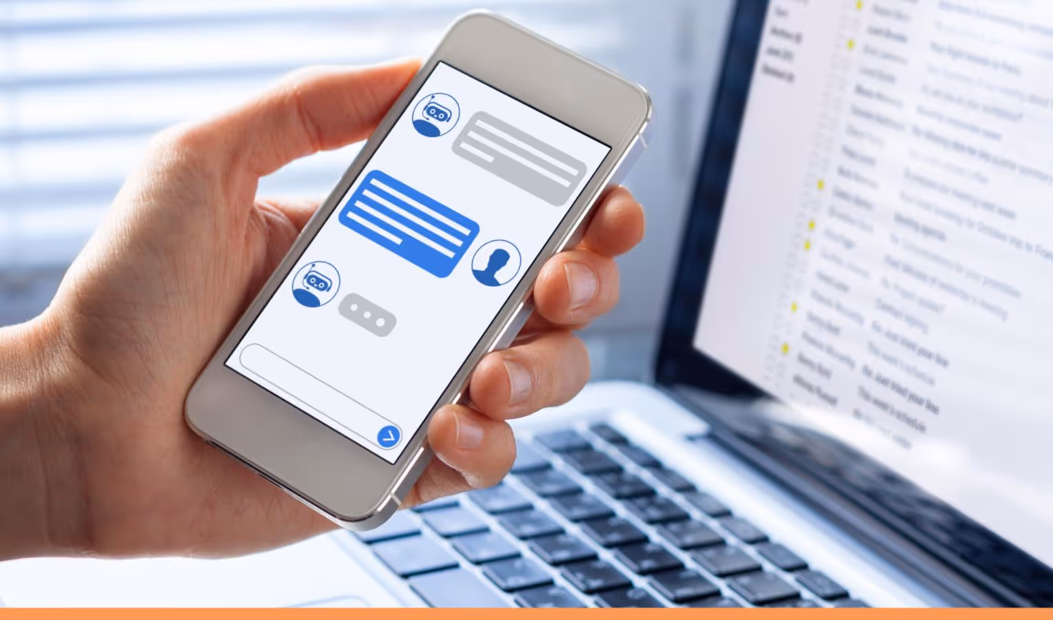 Hand hält ein Smartphone mit Chatbot-Konversation (blaue/graue Sprechblasen, Eingabefeld, Senden-Pfeil); im Hintergrund ein Laptop mit geöffnetem E-Mail-Posteingang.