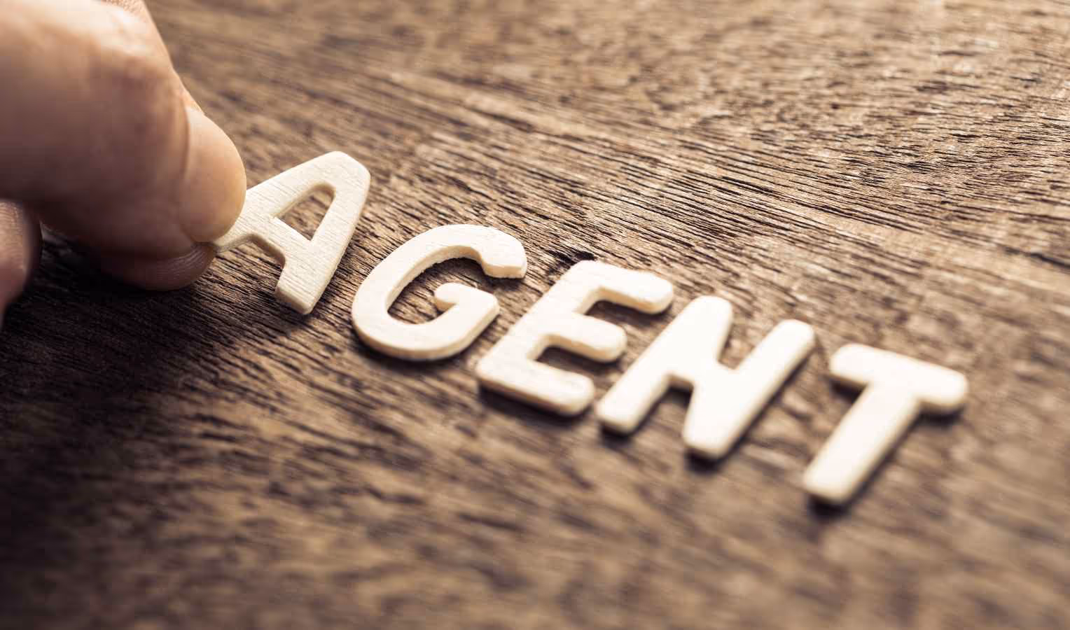Nahaufnahme: Eine Hand legt den Holzbuchstaben „A“ auf eine Holzoberfläche; daneben liegen weitere helle Buchstaben, die schräg das Wort „AGENT“ bilden (geringe Tiefenschärfe).