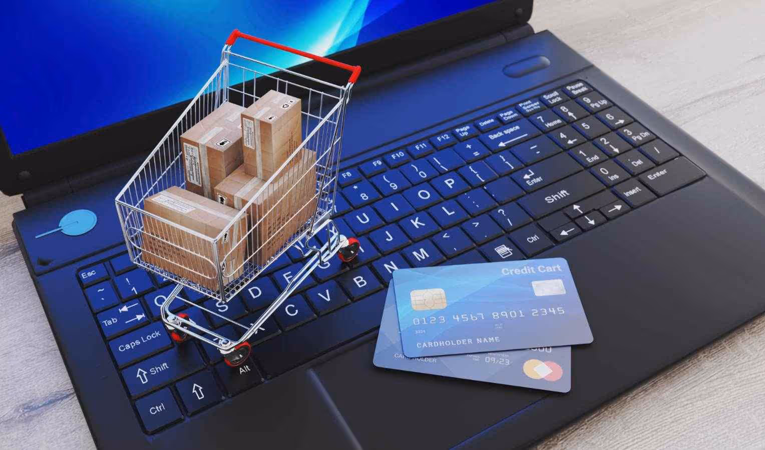 Miniatur-Einkaufswagen mit kleinen Versandkartons steht auf einer Laptop-Tastatur; daneben liegen zwei Kreditkarten – Symbol für Online-Shopping und Bezahlung.