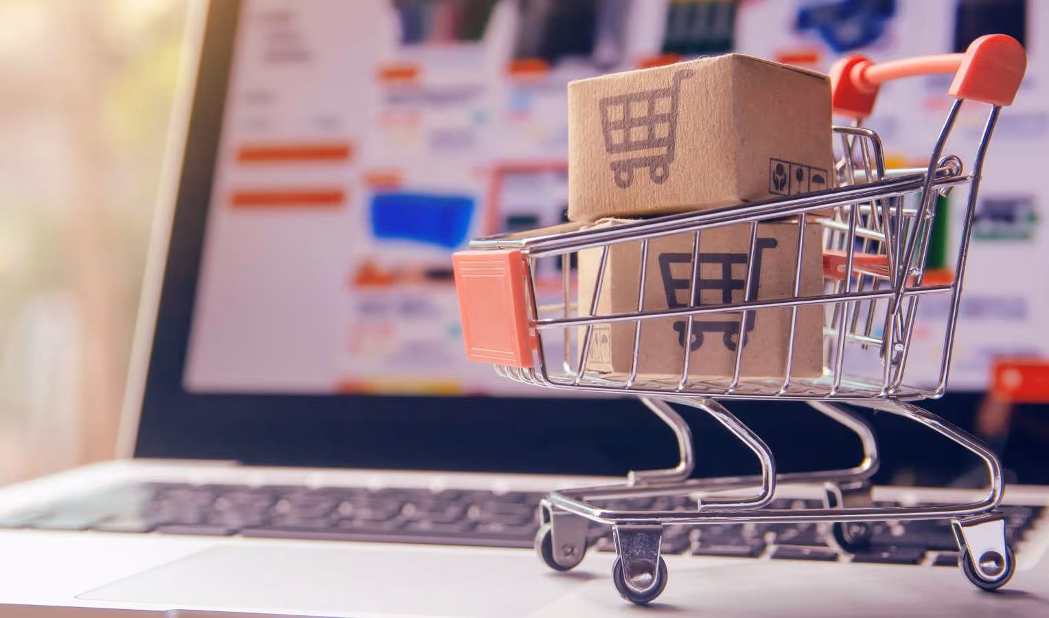 Miniatur-Einkaufswagen mit Paketen auf einer Laptop-Tastatur, unscharfer Online-Shop im Hintergrund (E-Commerce/Online-Bestellung).