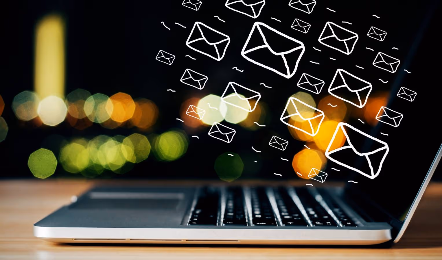 Seitlich fotografierter Laptop auf einem Tisch; im Vordergrund bunte Bokeh-Lichter, darüber fliegende gezeichnete Umschlag-Symbole (E-Mails).