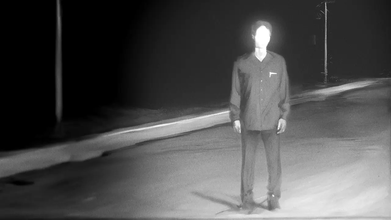 Faceless person standing alone on a dark, empty road at night, illuminated by a bright light, wearing a dark shirt and pants, with utility poles in the background.