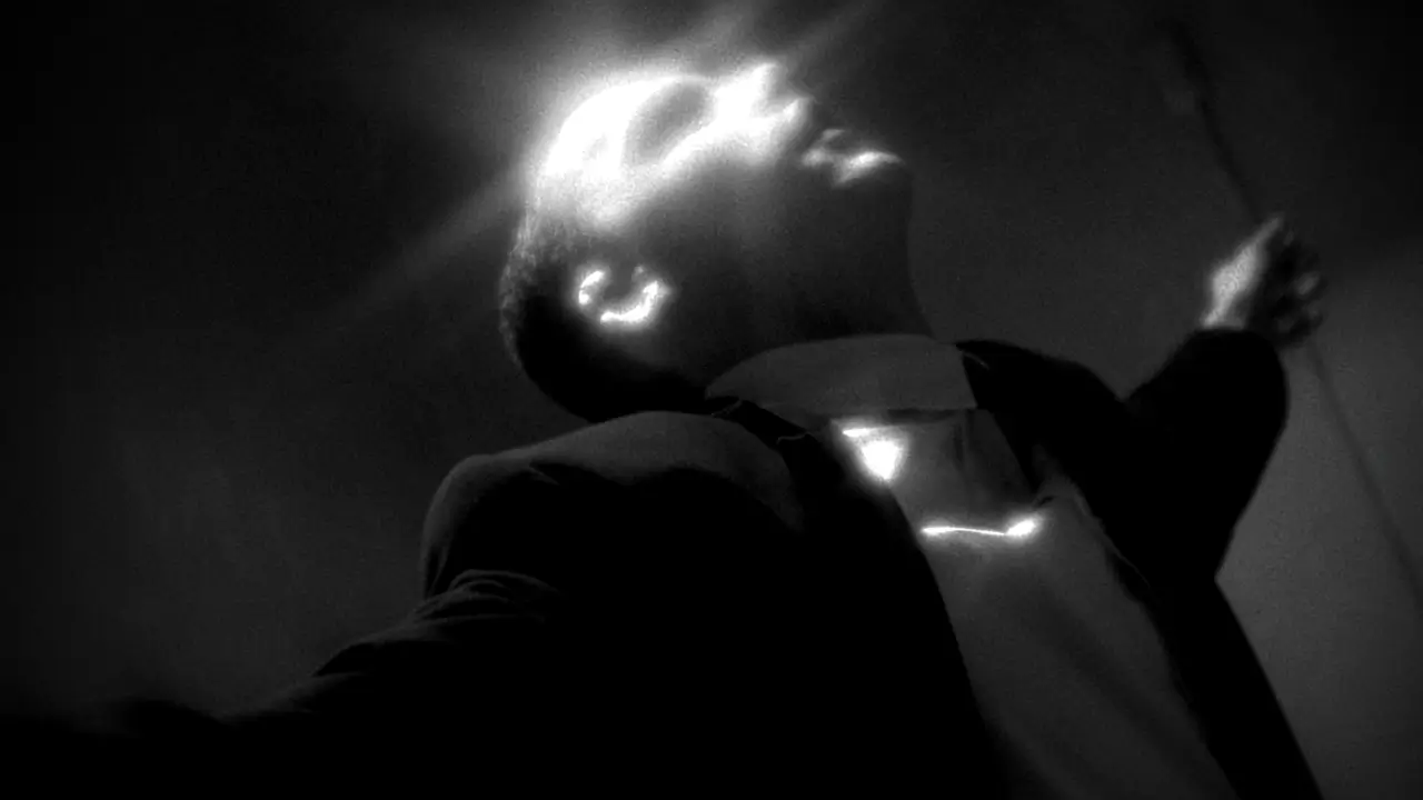 Black-and-white image of a man in a suit with arms outstretched and head tilted back, illuminated by dramatic light from above.