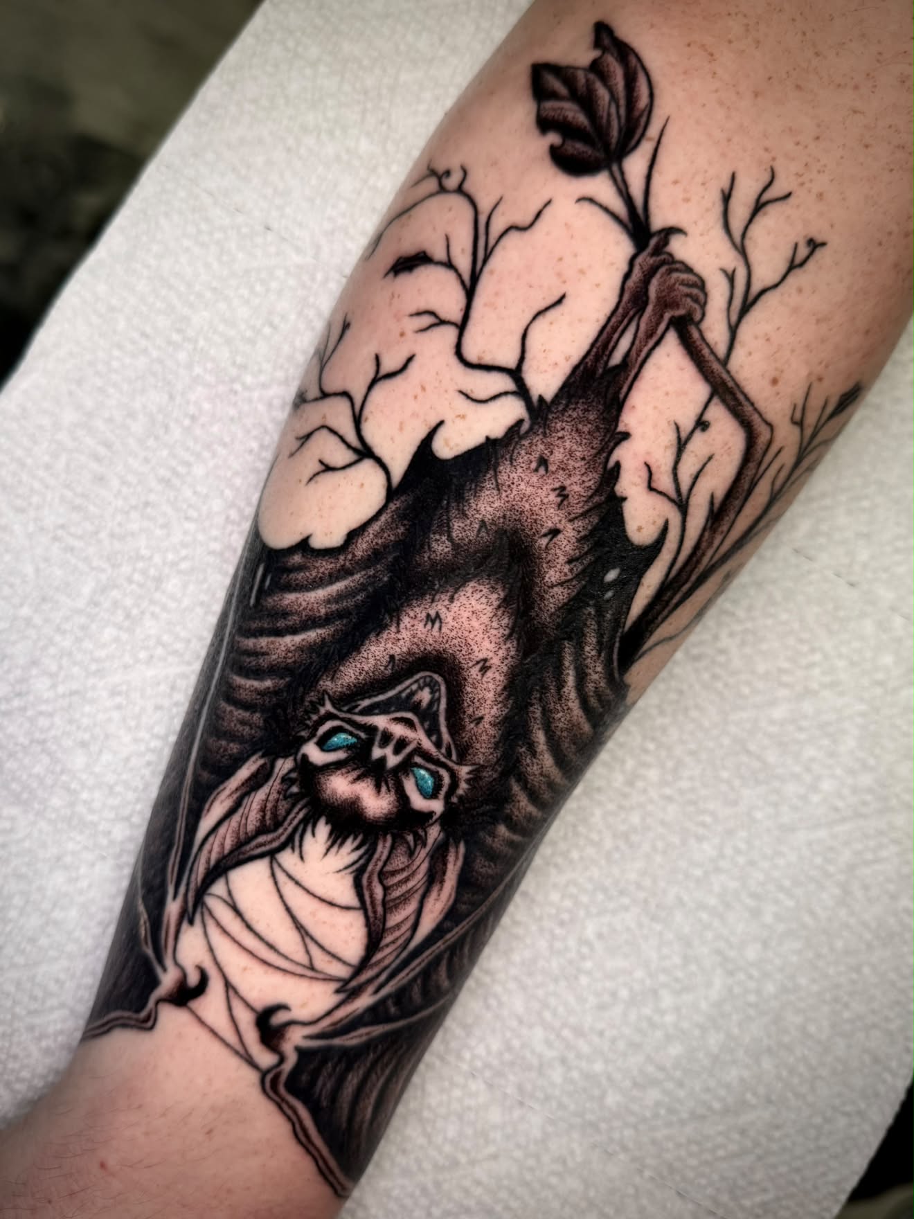 Blackwork bat hanging from a branch tattoo with blue eyes