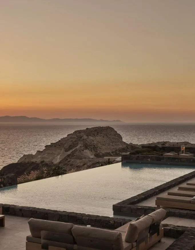 A swimmingpool on a greek island while the sunsets and the warm and orange glow of the sun touches the water and sky.