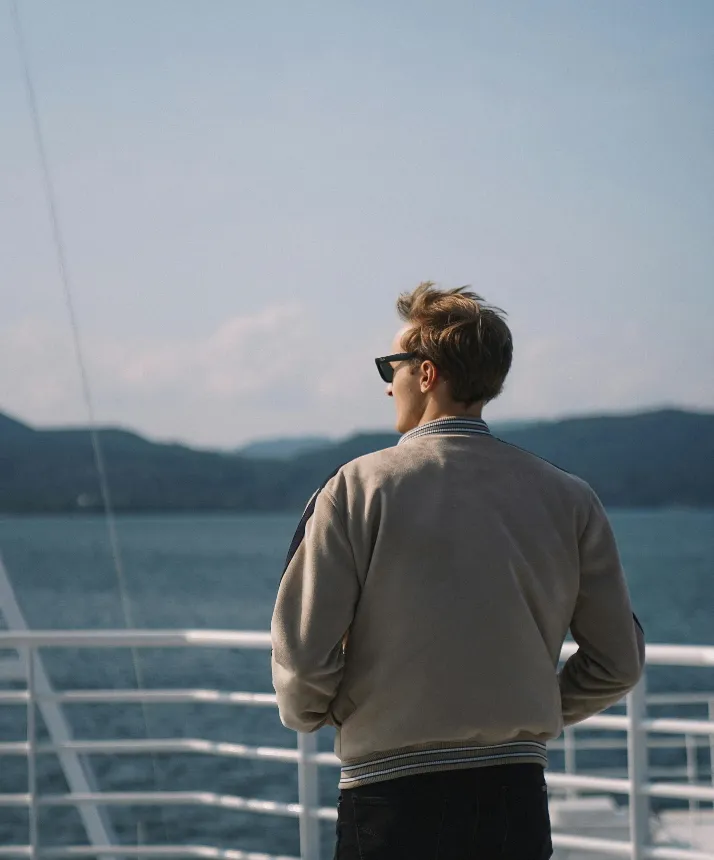 A guy standing in front of railing on a boat watching the Norwegian Fjords in the distance while wearing sunglasses.