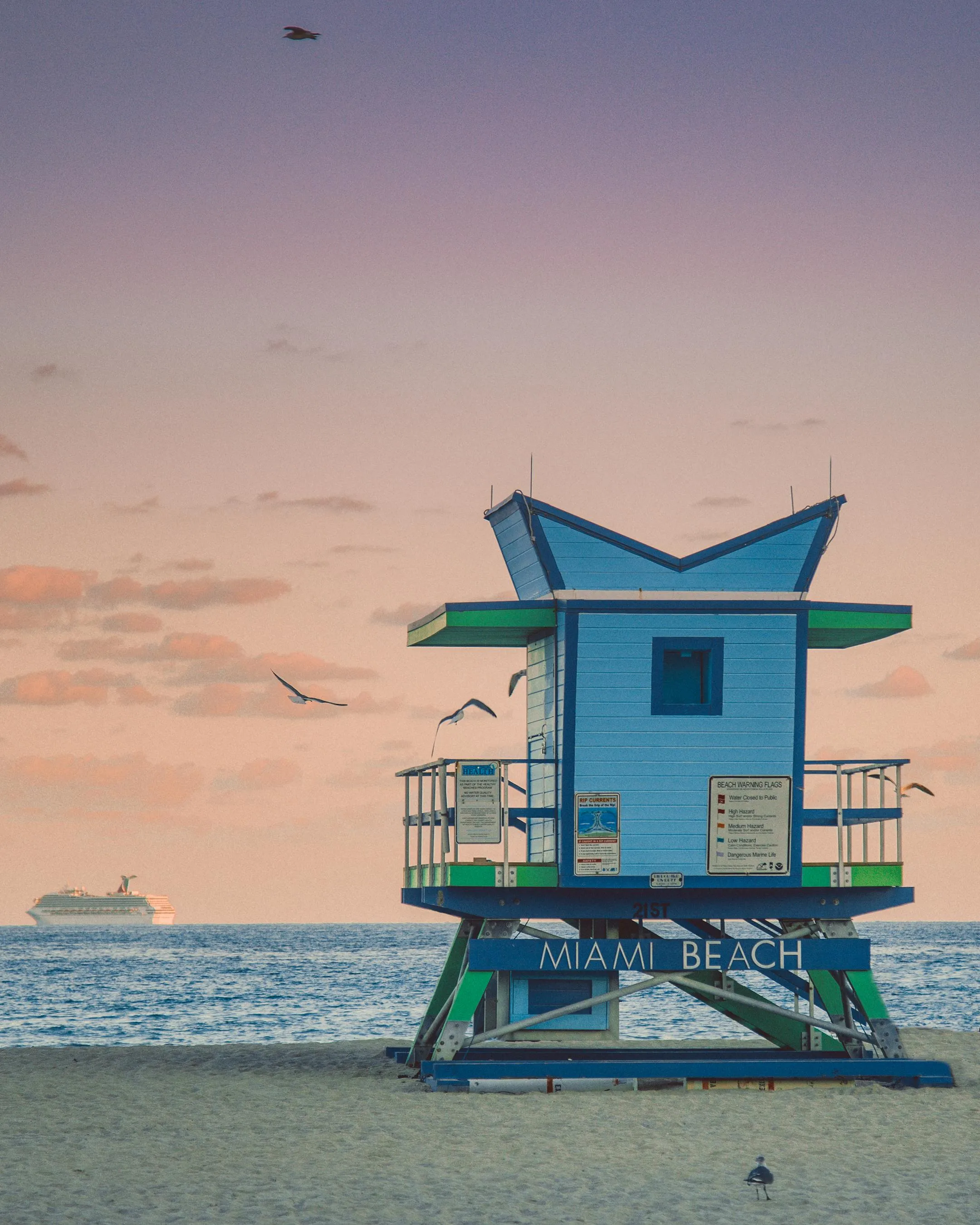 A colorful lifeguard tower at Miami Beach during sunset, with a cruise ship visible on the horizon and seagulls flying overhead.