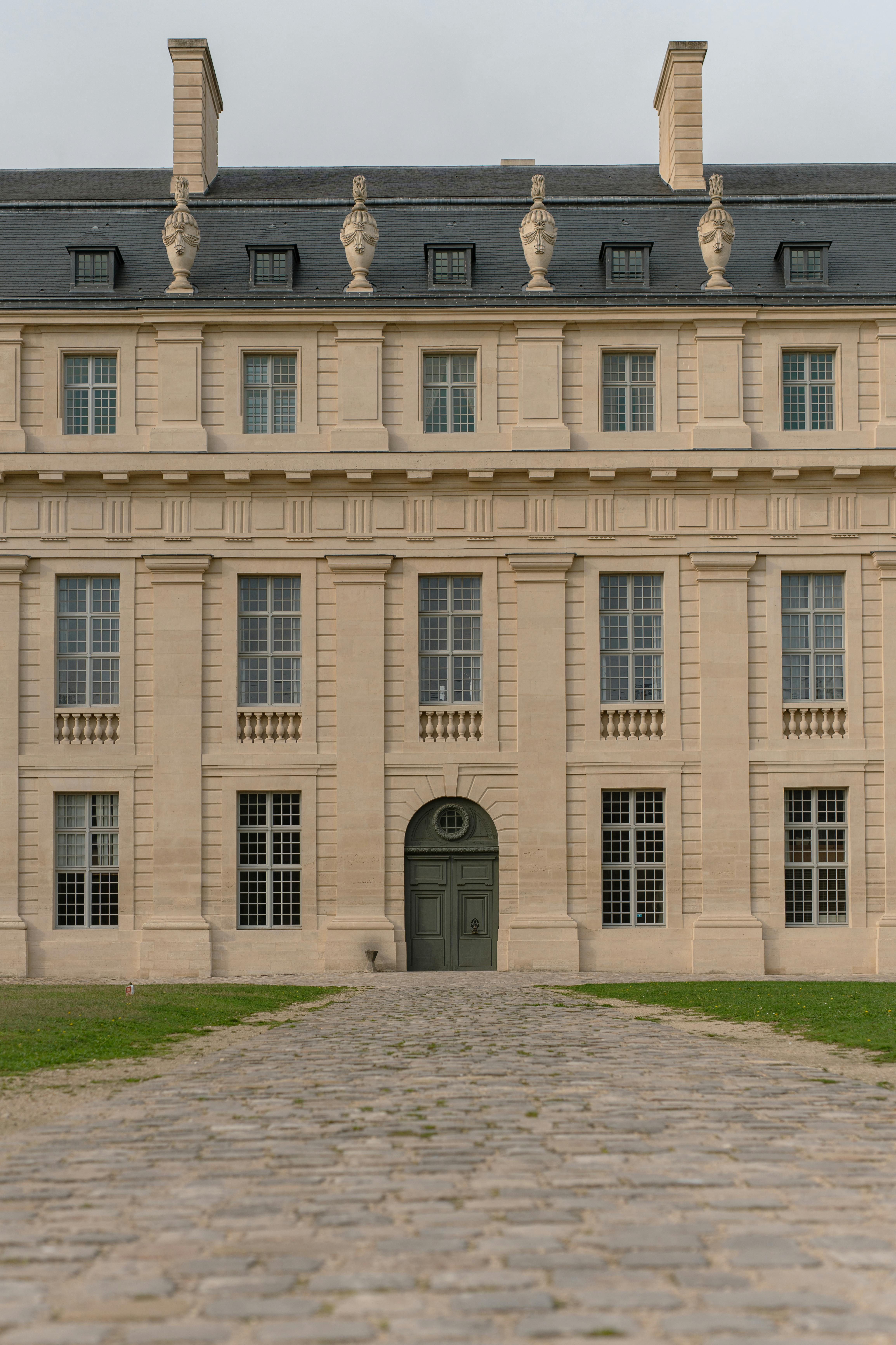 A grand, neoclassical building with a symmetrical facade featuring tall, narrow windows and ornate statues along the roofline. The entrance is marked by a large arched doorway, and the building is set back from a cobblestone pathway leading to the door. The structure exudes an air of historic elegance and architectural refinement.