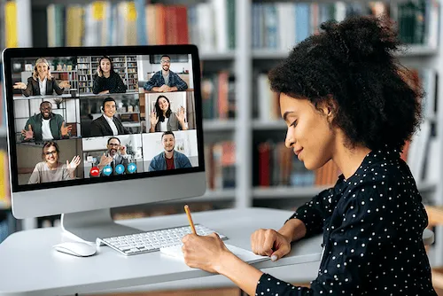 A woman sits in front of a virtual meeting, taking notes.