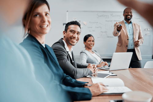 Smiling diverse colleagues sitting at a conference table with a man presenting in front of a whiteboard during a meeting.