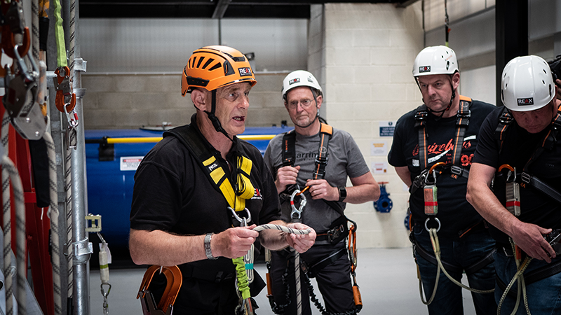 An instructor in an orange helmet suspended on a ladder structure demonstrates working at height techniques to a group of trainees in white helmets.