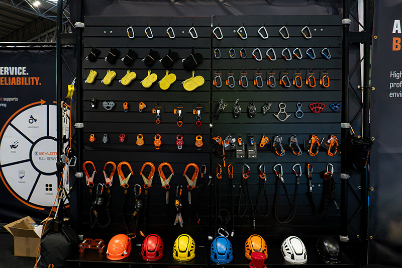 SKYLOTEC trade show pegboard displaying neatly organized safety equipment, including carabiners, pulleys, lanyards, and a row of colorful helmets.