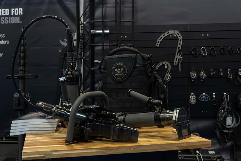 Close-up of heavy-duty black tactical rescue tools and a LIBERVIT B.A.S. backpack on a table, with dark carabiners on a pegboard behind them.