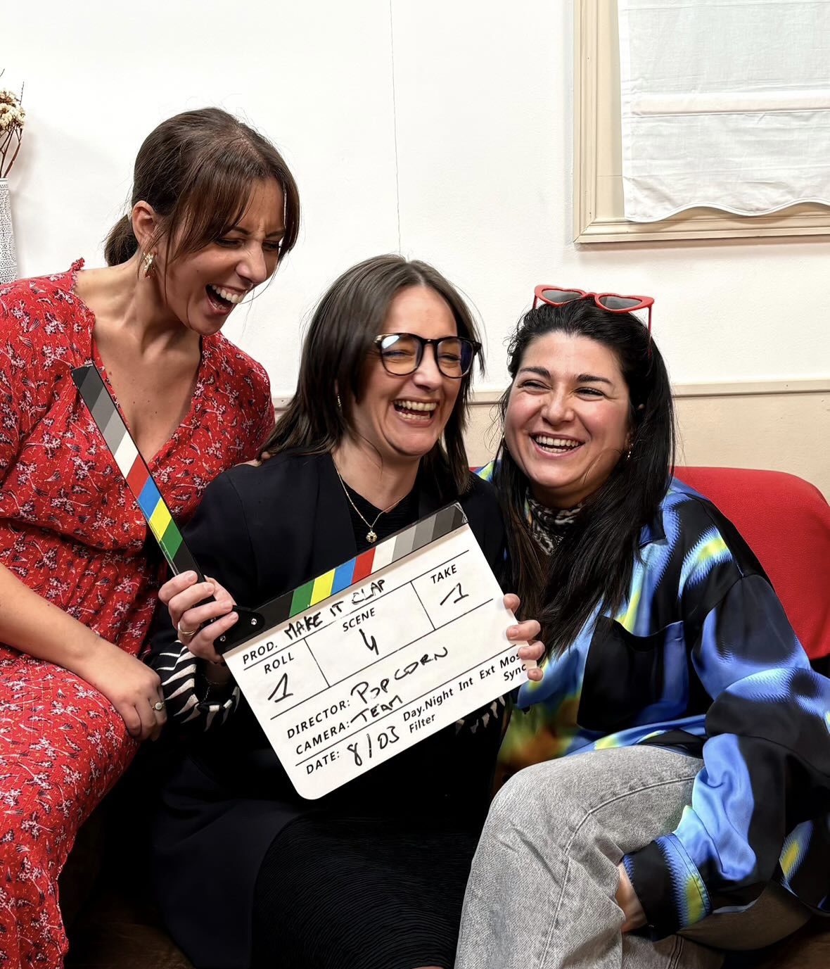 Three friends laughing on a couch holding a clapper board