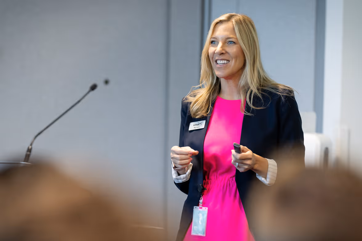 A Arthrex speaker in a pink blouse and black blazer addresses an audience in a conference room.
