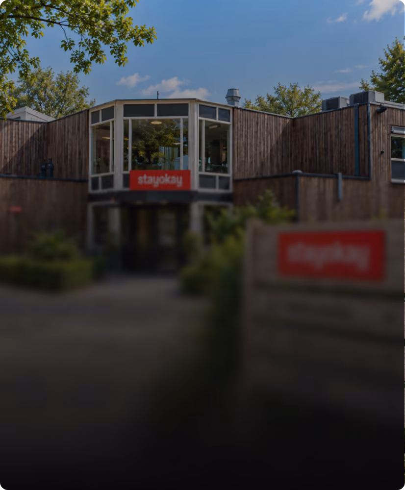 A modern building with wooden paneling and large windows under a blue sky, featuring red 'stayokay' signs on the front and a nearby wooden fence.